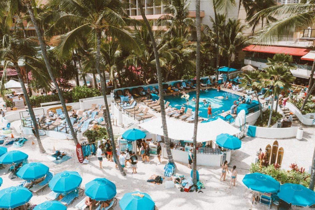 Photo of hotel guests swimming in the Outrigger Hotel pool surrounded by palm trees and overlooking beach loungers under umbrellas on Waikiki Beach