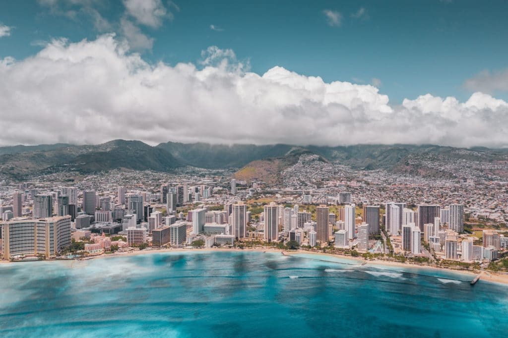 Aerial photo of Waikiki Beach. You can see rocks and coral through the bright blue water, hotels and buildings are lined behind the sand and continue off into the city. Mountains are in the background. The sky is blue with thick white clouds lingering low above the mountains.