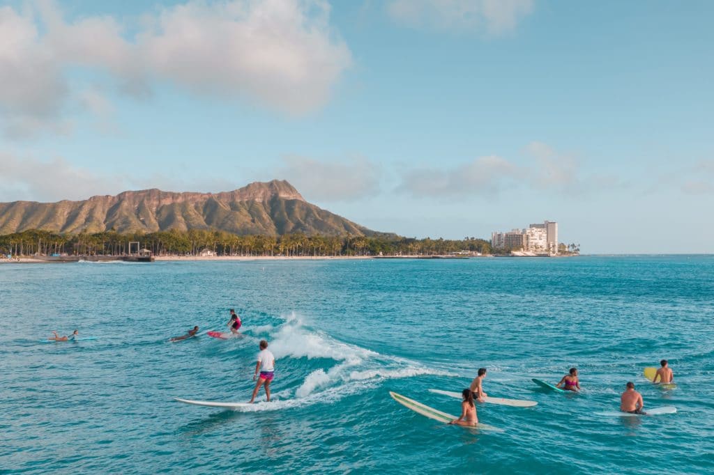 There are surfers out in the water on Waikiki Beach, catching waves on the bright blue water. Diamond Head is in the background and the sky is mostly clear with a few clouds.