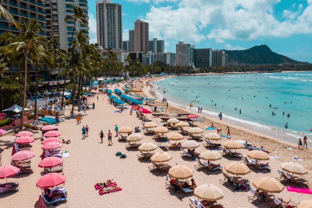 View of Waikiki Beach with building and palm trees lining the left-hand side. Beach umbrellas and lounge chairs are scattered throughout the sand and swimmers are in the bright blue ocean water. Diamond Head is in the background. The sky is blue with some light clouds.